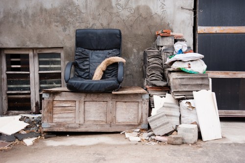 In an outdoor urban setting, a black office chair with a fabric seat and backrest is positioned on a weathered wooden chest or storage box. The chair's armrests are made of black plastic, and the cushion appears slightly worn. To the right of the chair, there is a pile of construction and renovation waste, including irregularly shaped concrete blocks with a rough surface, some with visible embedded aggregate, and a few white plasterboard pieces leaning against the blocks. The waste is stacked on a concrete pavement, with loose fragments and dust scattered around the base. Behind the chair and waste pile, a wall features a small ventilation grate made of horizontal metal slats set into a brick or concrete wall, with a section of black painted door or panel partially visible on the right side. The overall atmosphere suggests a typical rubbish clearance scene suitable for a commercial waste collection service operating in Bracknell Forest, with an emphasis on bulky waste and debris.