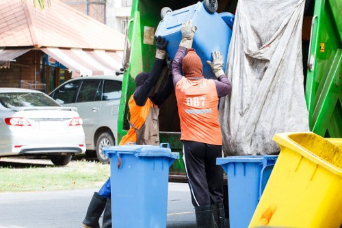 Two waste collection workers wearing high-visibility orange vests and protective gloves are lifting a large blue plastic bin into a green refuse collection vehicle parked on a residential street. The workers are positioned on either side of the bin, with one clearly visible and the other partially obscured, both focusing on loading the bin. The vehicle has an open compartment at the rear where a grey tarp covers part of the waste area. In the foreground, there are additional bins: a yellow bin on the right and two blue bins on the left, all placed on the pavement. In the background, there are parked cars, including a white vehicle, along with elements of a suburban environment such as a house with a red-tiled roof, a tree with green leaves, and a part of a street scene indicating a typical residential neighbourhood, possibly within the Bracknell Forest area. The scene is well-lit with natural daylight, highlighting the setup for rubbish collection services in the local area. Commercial Waste Bracknell Forest’s focus on waste management is reflected through the activity, emphasizing the routine of rubbish removal in this community.