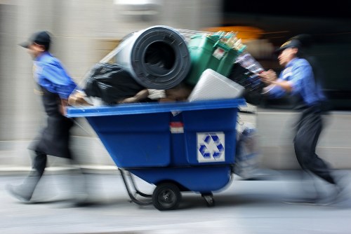 A blue recycling bin mounted on a wheeled cart is overflowing with various waste items including a large black plastic bag, a green wheelie bin, and other miscellaneous rubbish. The bin is situated outdoors, likely on a driveway or street in Bracknell Forest, with three individuals pushing the bin from opposite sides, appearing to be in motion. The waste materials extend beyond the rim of the bin, indicating a collection or disposal activity associated with commercial or domestic rubbish removal services. The background shows a blurred urban environment with concrete surfaces, and the scene is illuminated by natural daylight, emphasizing the textures of the waste and the surrounding area. This image relates to rubbish collection and waste disposal services offered by Commercial Waste Bracknell Forest in the local area.