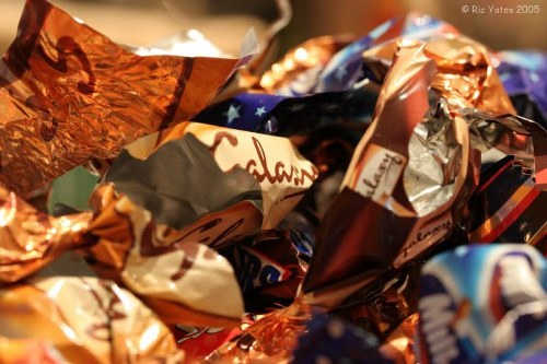 A close-up image of a pile of crumpled and twisted foil wrappers in various metallic colours, including shades of copper, silver, and blue, with some featuring printed branding or patterns. The wrappers appear to be made of shiny, reflective materials, and are stacked chaotically, occupying most of the foreground. In the background, there is a hint of a blurred surface, possibly a table or countertop, with warm ambient lighting that highlights the gloss and creases of the wrappers. The overall scene suggests an indoor setting, potentially near a recycling or waste collection area, and is representative of discarded confectionery wrappers often collected by rubbish removal services such as Commercial Waste Bracknell Forest, serving local areas including Bracknell and surrounding postcode zones. The image emphasizes the texture and metallic finish typical of packaging waste awaiting disposal or recycling as part of rubbish collection activities.