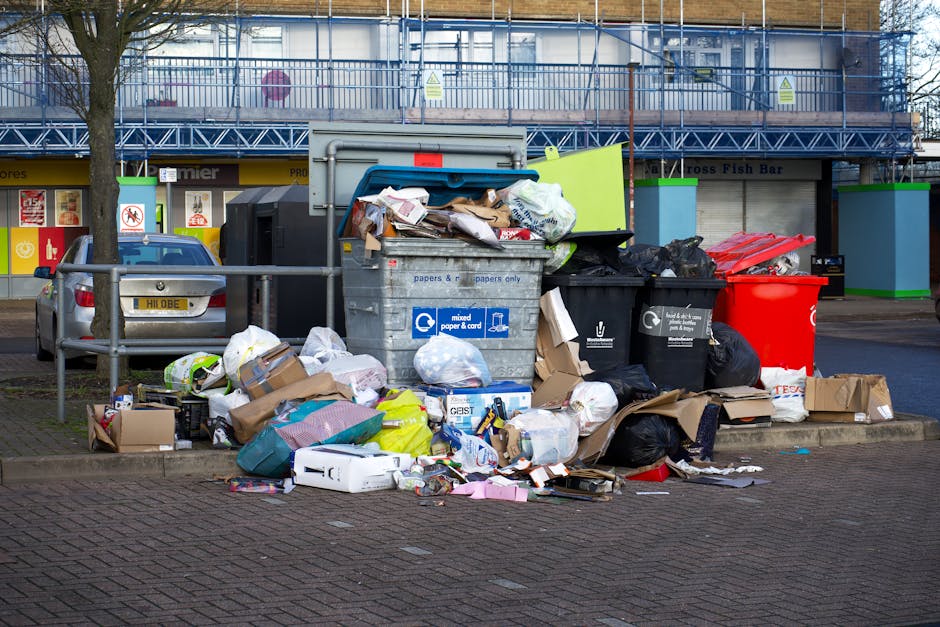 A close-up view of a roadside waste collection point featuring a pile of mixed rubbish, including black plastic garbage bags, cardboard boxes, and discarded household items such as cushions with fabri