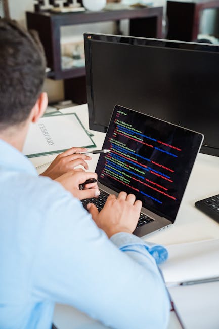 A person wearing a light blue shirt is seated at a desk, working on a laptop that displays lines of colorful computer code on its screen. The individual is holding a wireless mouse with one hand and a